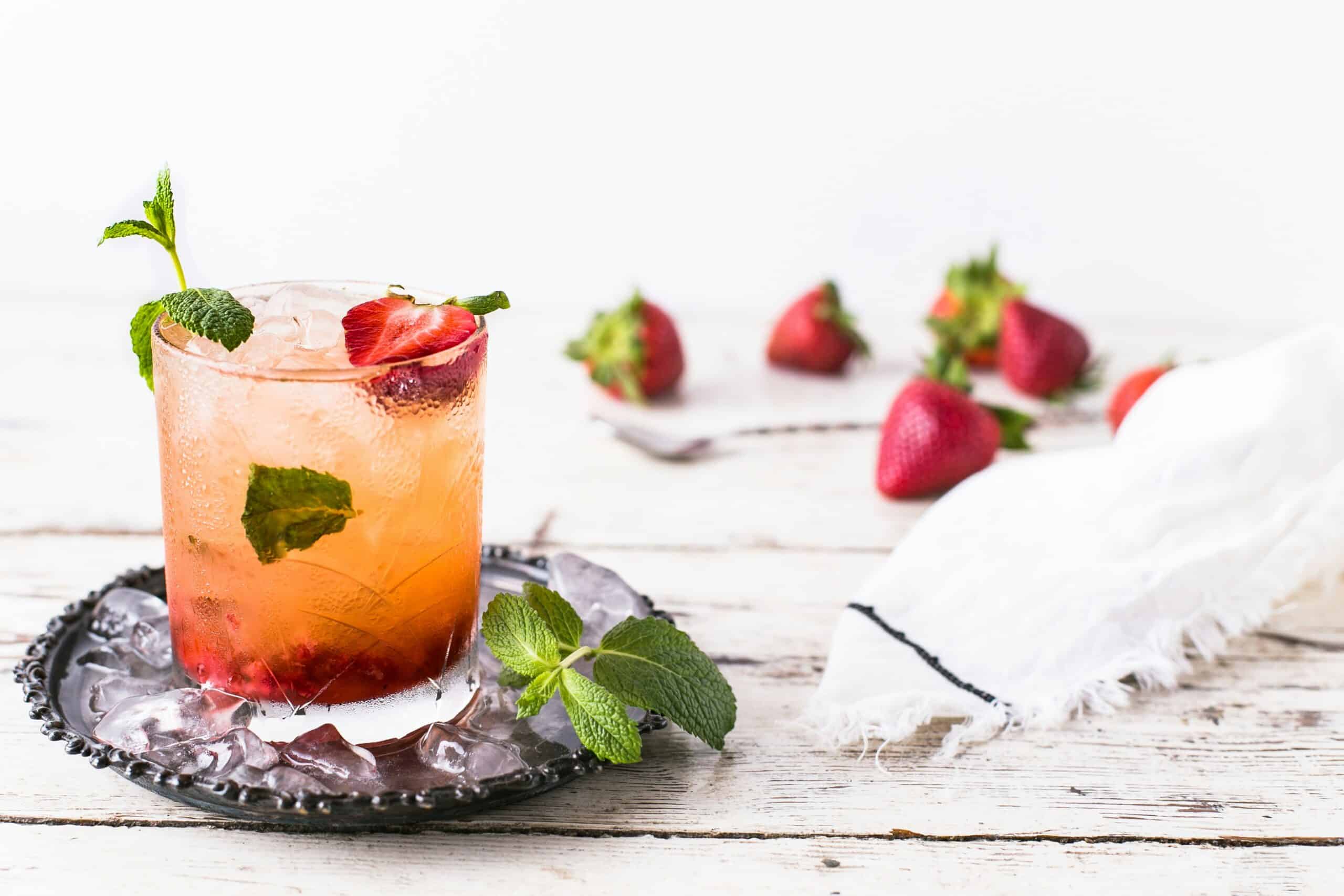 Rocks glass with strawberry mint mocktail on a black plate with whole strawberries scattered around