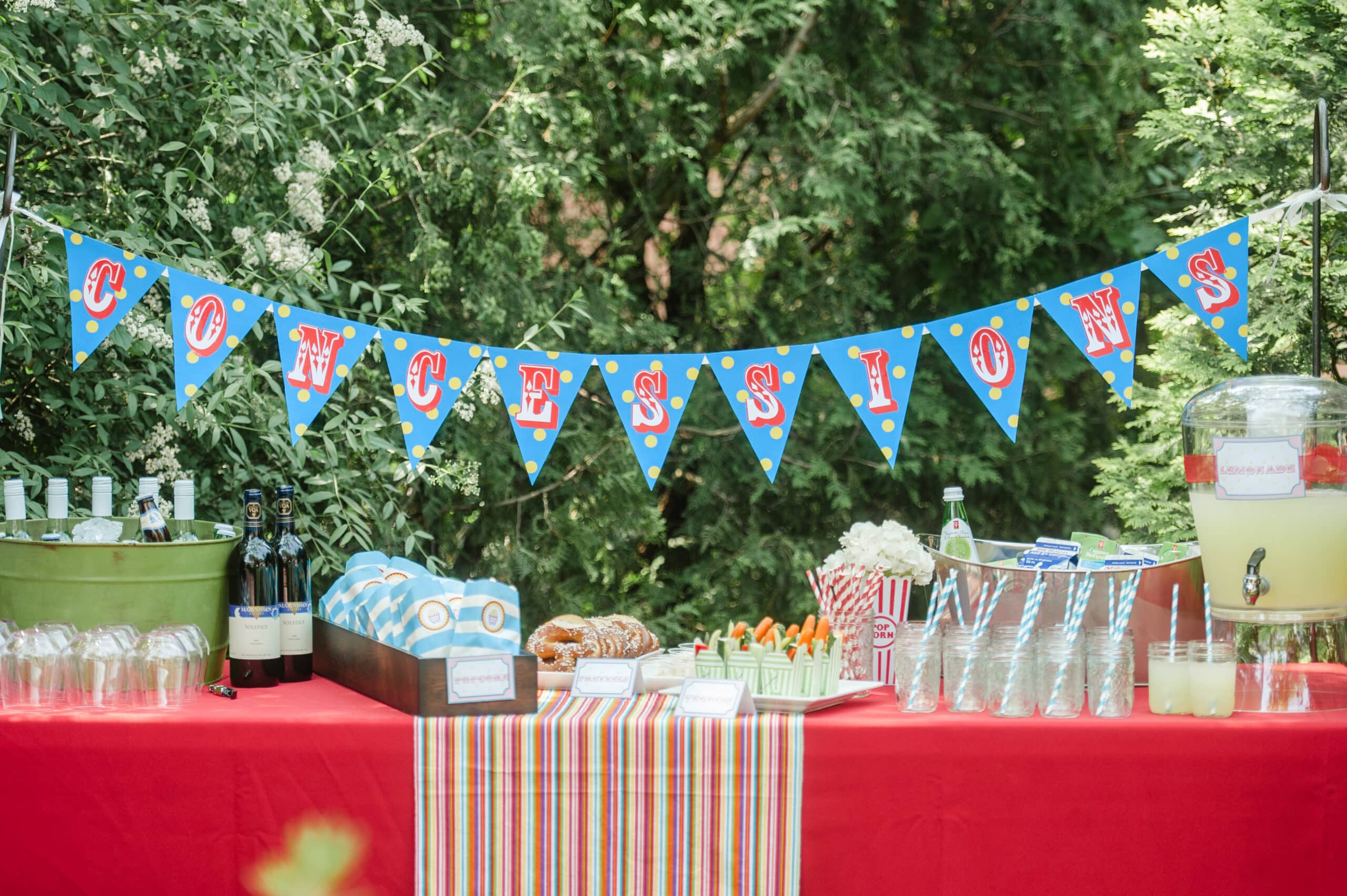 Concession stand at circus-themed birthday party with beverages, bags of popcorn, mini take out boxes of crudites, lemonade, candies on colourful linen