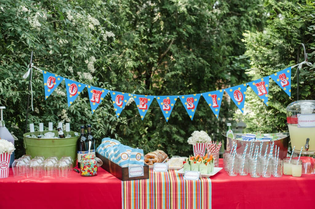 Concession stand at circus-themed birthday party with beverages, bags of popcorn, mini take out boxes of crudites, lemonade, candies on colourful linen